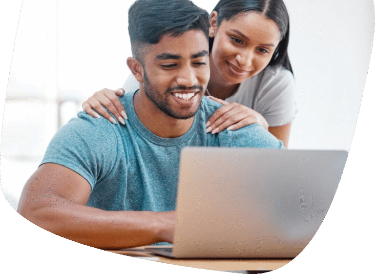 A smiling couple looks at a laptop together. The man is seated and typing while the woman stands behind him, resting her hands on his shoulders, both appearing happy and engaged with the screen.