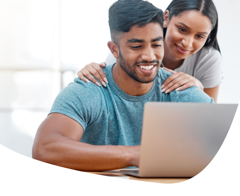 A smiling man sits at a desk using a laptop while a woman stands behind him, resting her hands on his shoulders and looking at the screen. Both appear happy and focused.