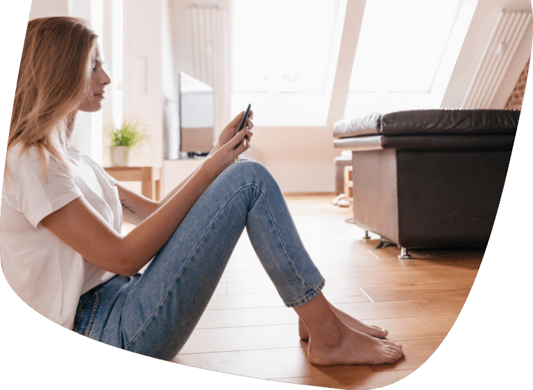 A woman sits barefoot on a wooden floor, leaning against a wall and looking at her phone. She wears a white t-shirt and blue jeans. The room has large windows, a sofa, and a potted plant in the background.