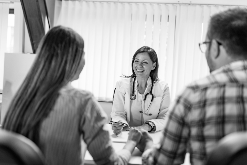 A female doctor smiles while speaking with a couple sitting across from her in an office. The doctor holds a pen and notepad, and the couple holds hands, suggesting a supportive consultation.