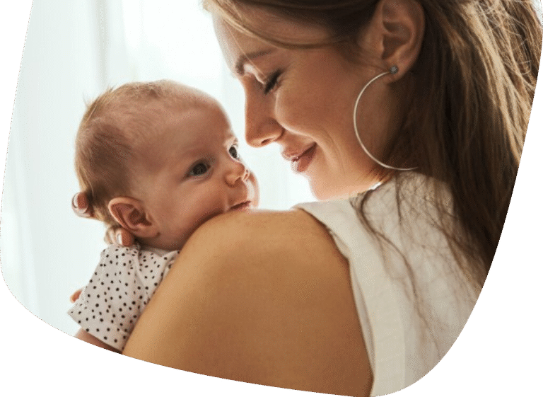 A woman with long brown hair smiles gently, holding a baby with light hair on her shoulder. The baby looks up, wearing a white outfit with black dots. Both appear calm and content in soft, natural light.