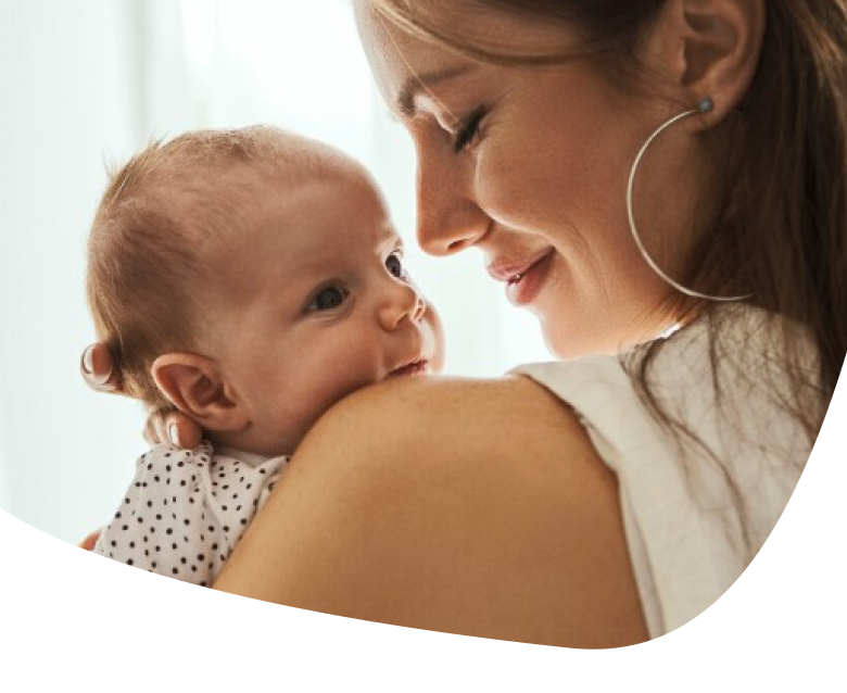 A woman smiles gently while holding a baby close to her shoulder. The baby looks up at her with a calm expression. Both are indoors with soft, natural light.