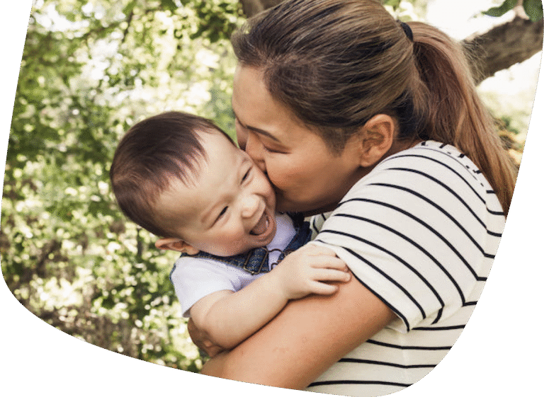 A woman hugs and kisses a smiling baby outdoors, surrounded by greenery. Both appear happy and affectionate.