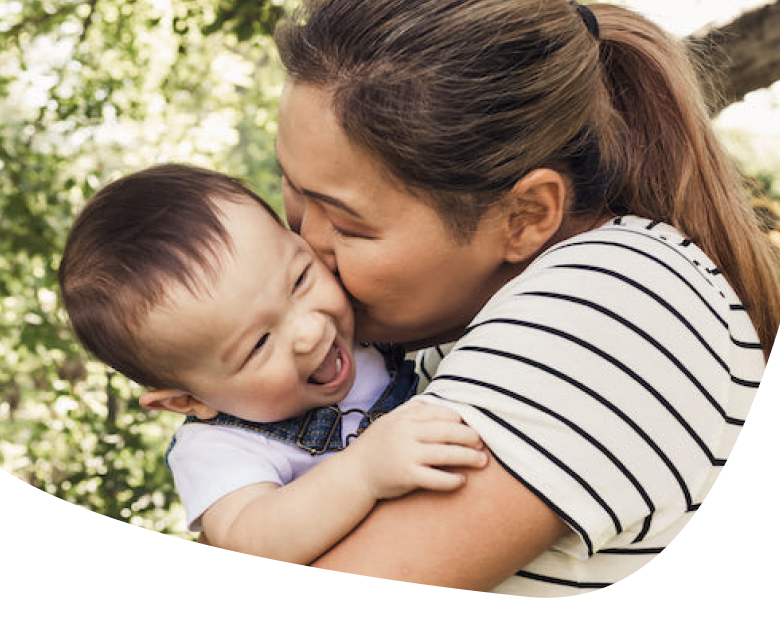 A woman with brown hair in a ponytail, wearing a striped shirt, holds and kisses a smiling baby outdoors. The baby looks happy and is wearing a white shirt with denim overalls. Green trees are in the background.