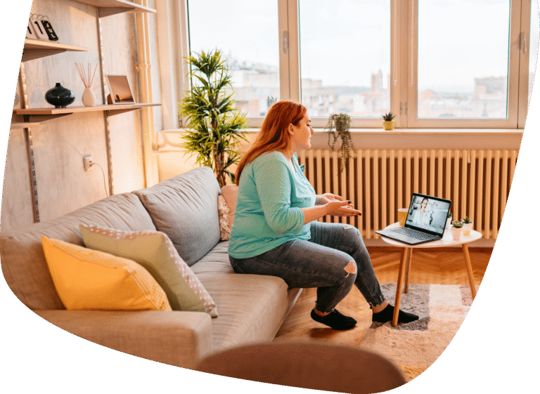 A woman sits on a gray couch in a cozy living room, having a video call on her laptop, which is placed on a small round table beside her. She gestures with her hands while talking.