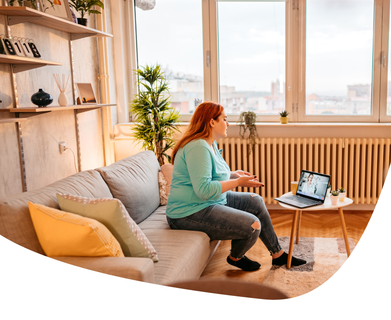 A woman sits on a sofa in a cozy living room, gesturing while having a video call on her laptop, which is placed on a small table in front of her. The room has large windows and various decorations.