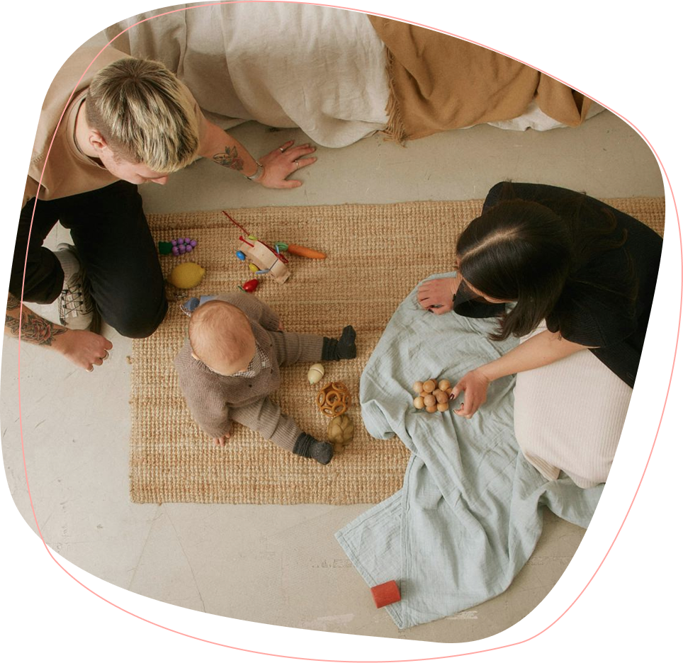 Two adults and a baby sit on a woven rug with toys scattered around. The adults face the baby, who is sitting up and playing, while one adult holds small wooden objects. The scene appears warm and relaxed.
