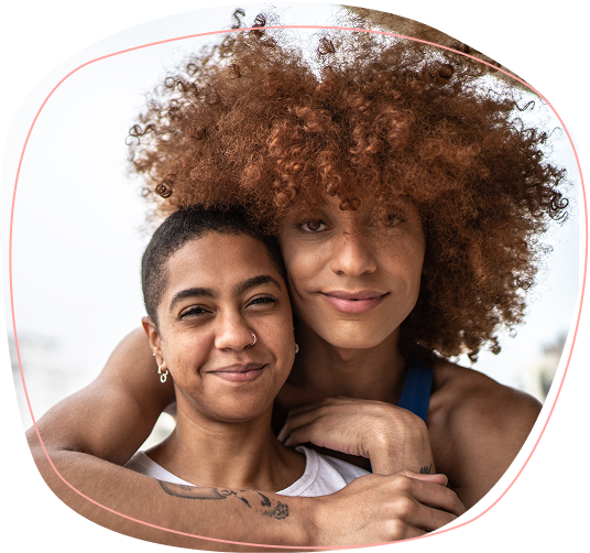 Two people smile while embracing each other closely. One person has short dark hair and a nose ring, while the other has voluminous curly hair and lightly rests their chin on their friends head. Both appear happy and relaxed.