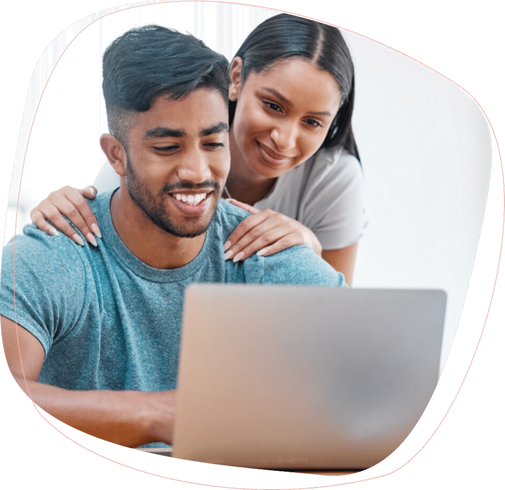 A smiling man sits at a laptop while a woman stands behind him, gently resting her hands on his shoulders and looking at the screen. Both appear happy and focused.