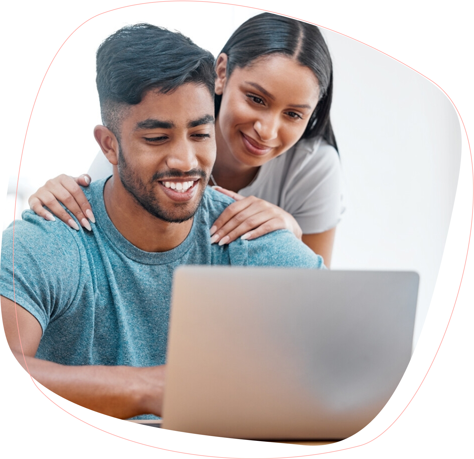 A smiling man sits at a laptop while a woman stands behind him, gently resting her hands on his shoulders and looking at the screen. Both appear happy and focused.