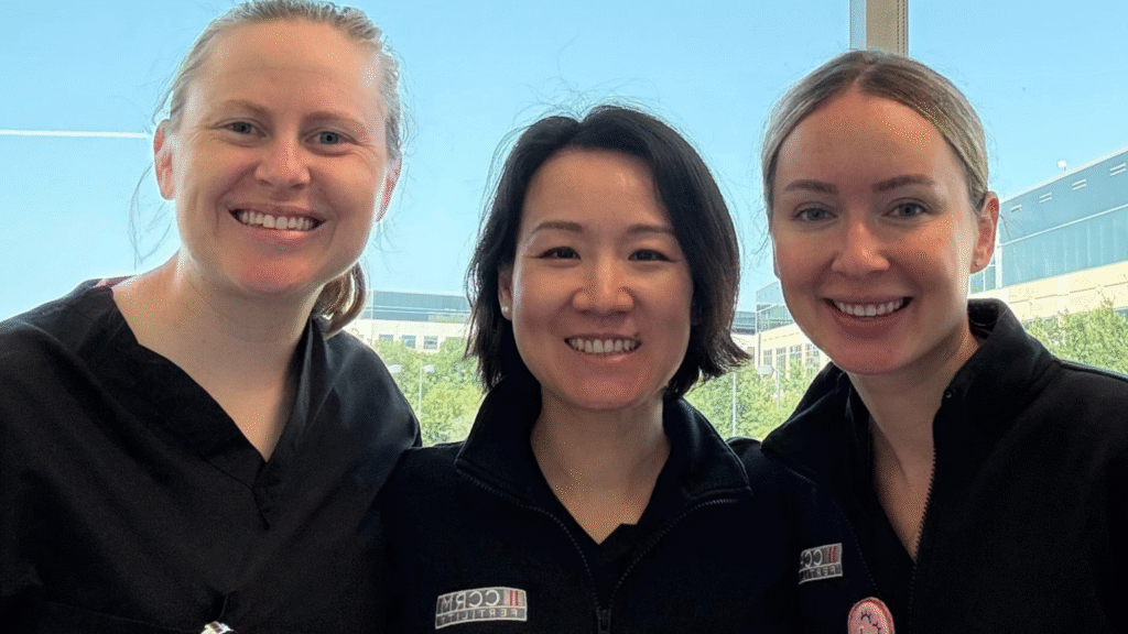 Three women in black uniforms smile together indoors with large windows behind them showing a bright blue sky and buildings outside.