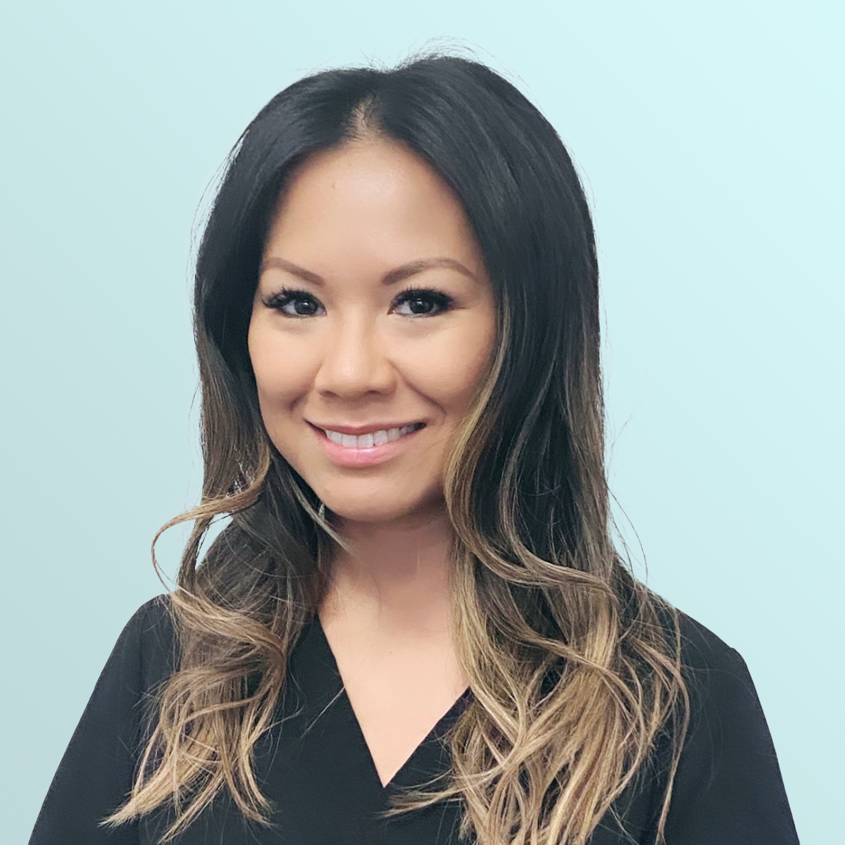 A woman with long, wavy dark hair and light highlights smiles at the camera. She is wearing a black top and is standing in front of a light blue background.