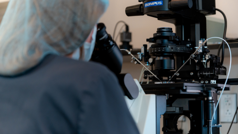 A person wearing a hair cover and lab coat looks through a microscope in a laboratory setting, focusing on scientific equipment with wires and mechanical components.