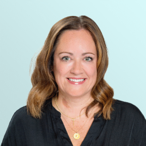 Smiling woman with shoulder-length light brown hair, wearing a black blouse and gold necklaces, posing in front of a light blue background.