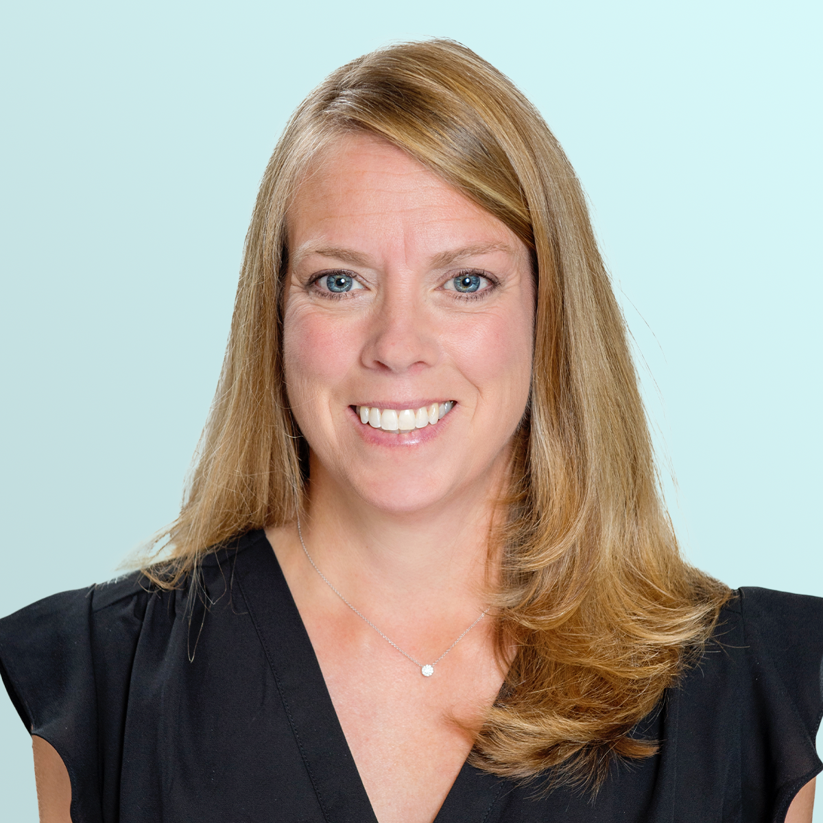 A woman with long blonde hair and blue eyes smiles at the camera. She is wearing a black top and a small pendant necklace, set against a light blue background.