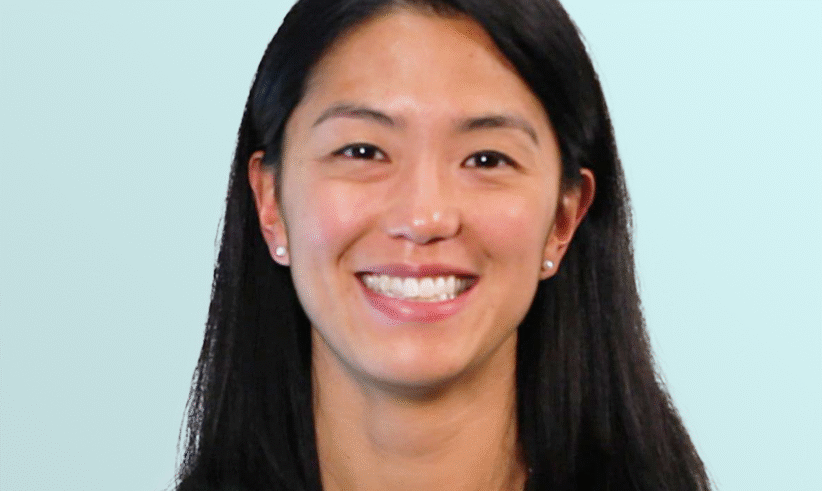 A woman with straight black hair and a floral necklace, wearing a sleeveless black top, smiles in front of a light blue background.
