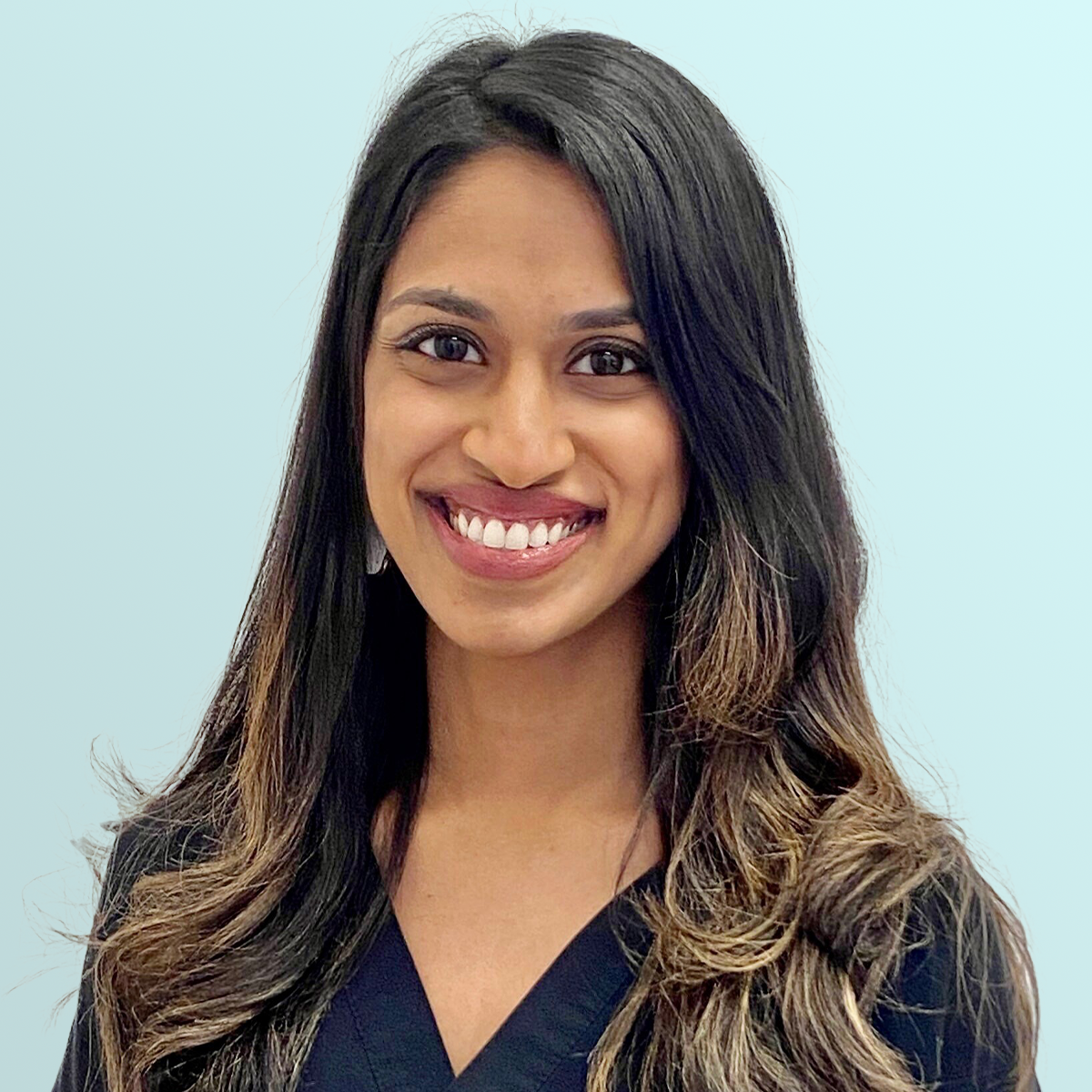 A woman with long, wavy dark hair wearing a black top smiles at the camera against a plain light blue background.