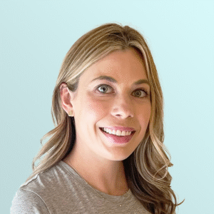 A woman with long, light brown hair and hazel eyes smiles at the camera. She is wearing a grey top and small earrings, with a light blue background behind her.