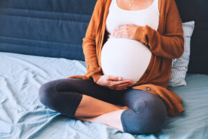 A pregnant person sits cross-legged on a bed, wearing a white top, brown cardigan, and gray leggings, with hands resting on their belly.