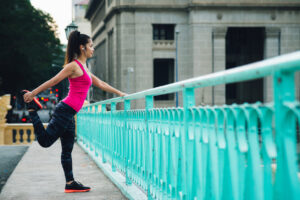 A woman in athletic clothing stretches her leg on a turquoise railing of a bridge, listening to music with earphones, with city buildings in the background.