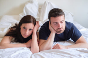 A man and woman lie side by side on a bed with white bedding, both resting their heads on their hands and looking serious or thoughtful. They appear to be lost in thought or concerned.