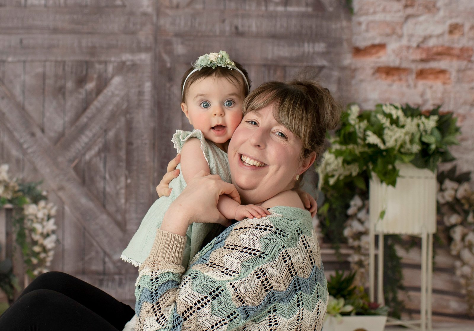 A smiling woman holds a baby girl dressed in pale green with a floral headband. They are seated in front of a rustic backdrop with flowers and wooden textures. Both look happy and close together.