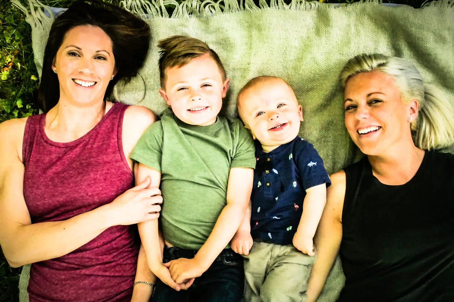 Two women and two young boys lie on a blanket outdoors, smiling at the camera. The women are on each end, with the children between them, creating a happy, relaxed family scene.