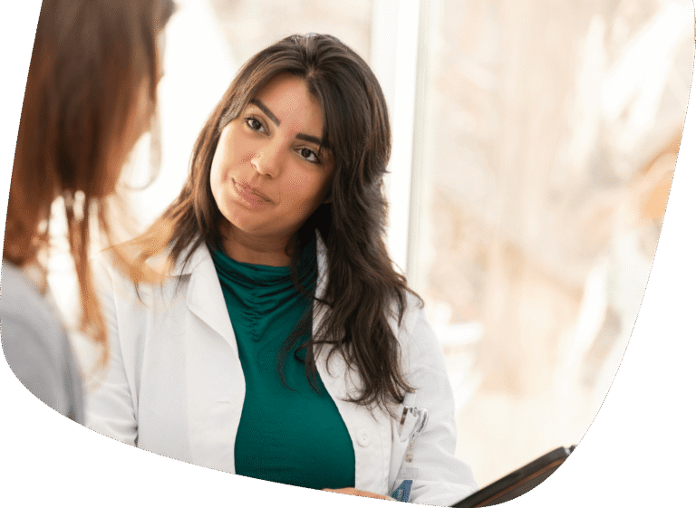 A female doctor in a white coat attentively listens to a patient during a consultation in a bright office setting.