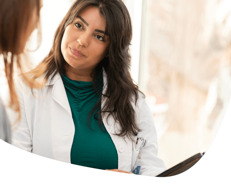 A woman wearing a white lab coat and green top attentively listens to another person during a conversation in a bright indoor setting.