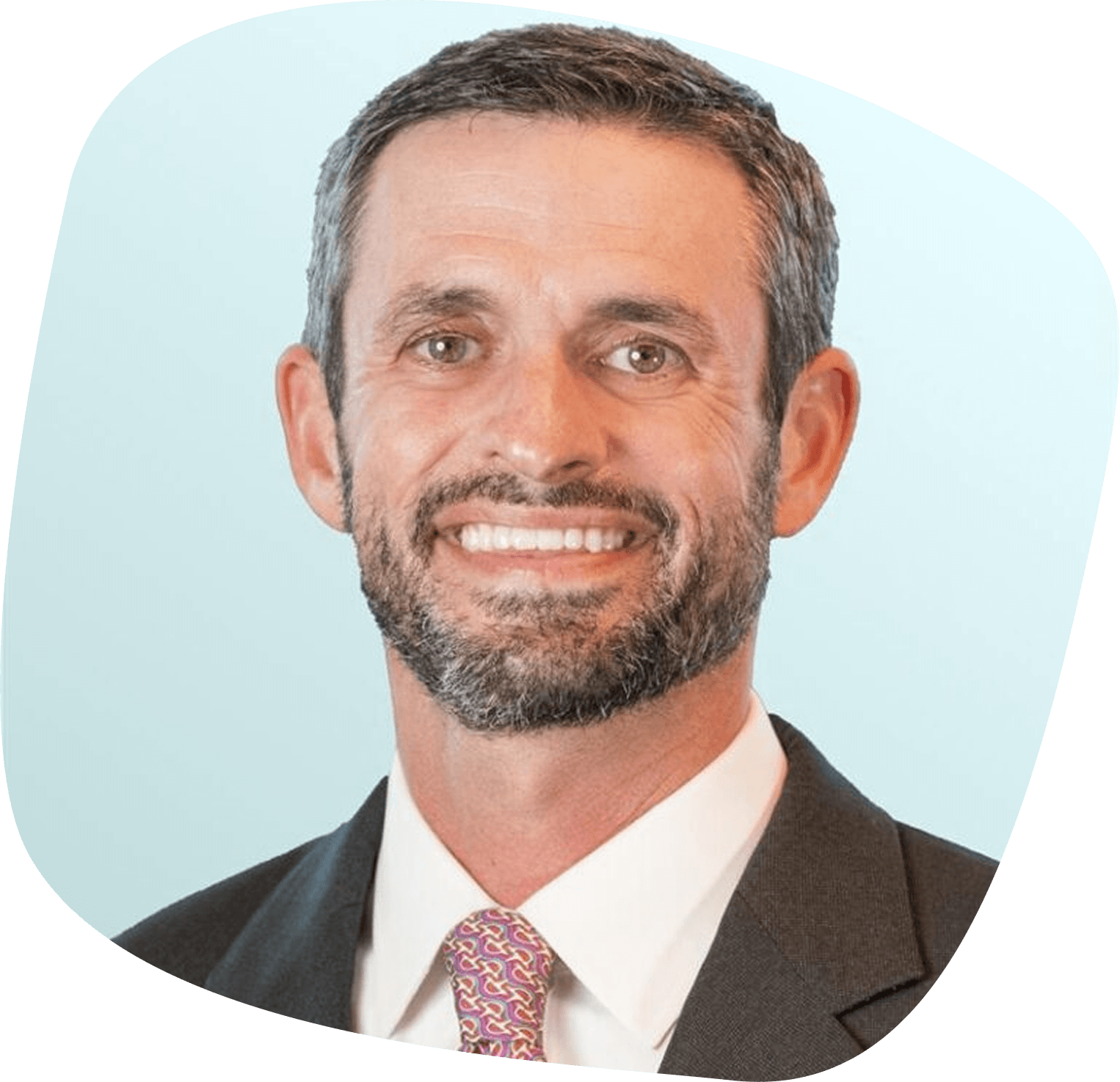 Smiling man with short dark hair and a beard, wearing a dark suit, white shirt, and patterned purple tie, in front of a light blue background.