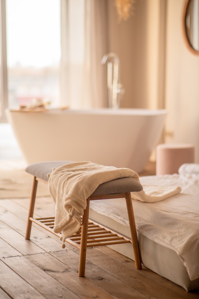 A cozy bathroom scene with a freestanding white bathtub in the background, perfect for a relaxing baking soda bath to support fertility, a soft towel on a cushioned wooden bench, and warm sunlight streaming through the window onto wooden floors.