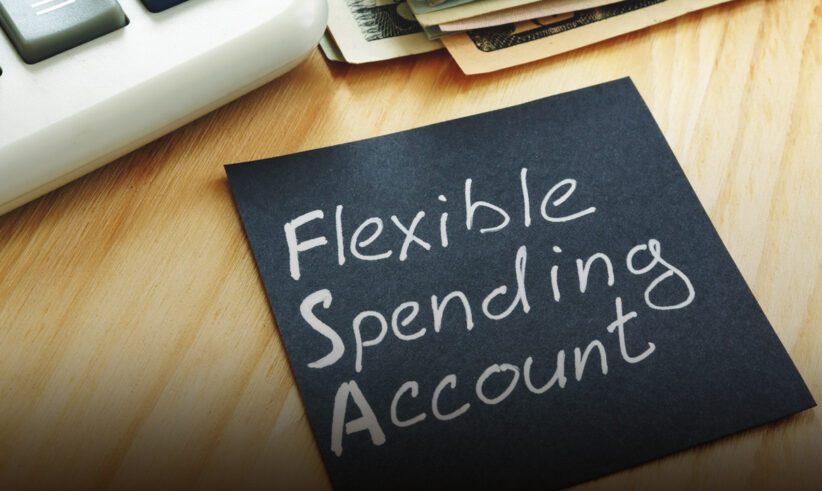A black sticky note with Flexible Spending Account written on it sits on a wooden desk near a calculator and a stack of cash.