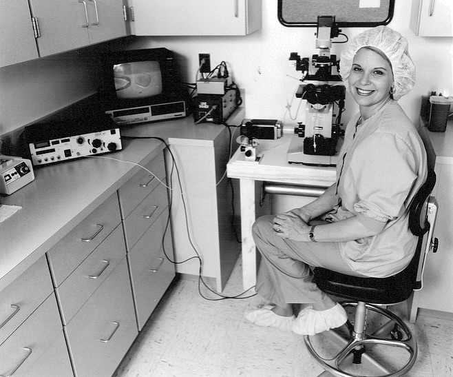 A woman in scrubs, a hair covering, and shoe covers sits on a stool beside a microscope and lab equipment in a laboratory, smiling at the camera.