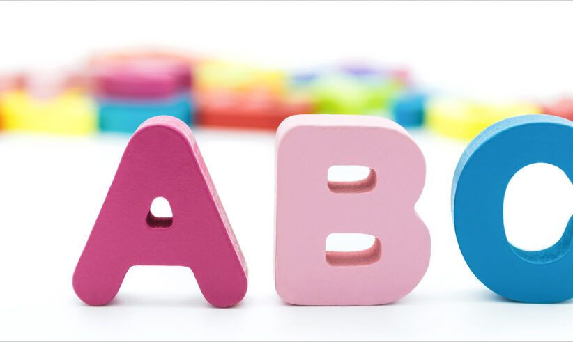 Three colorful wooden letters—A in red, B in pink, and C in blue—stand upright in the foreground, with blurred multicolored shapes in the background on a white surface.