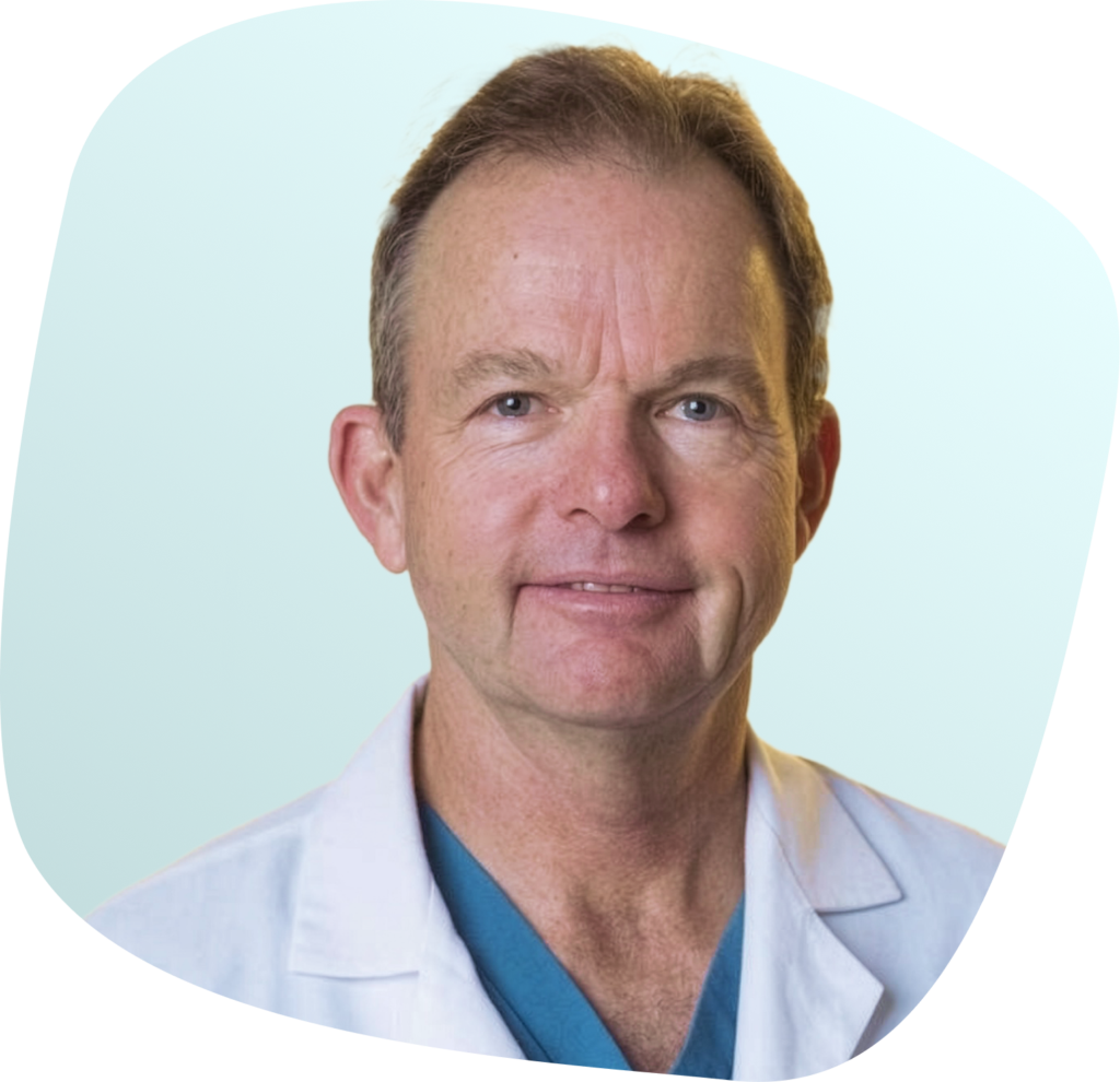 A middle-aged man with short light brown hair, wearing a white lab coat and a teal scrub top, smiles slightly against a light blue background.