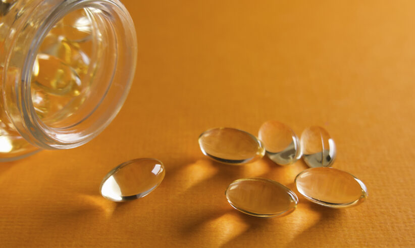 A glass bottle tipped on its side spills several oval, translucent, yellow softgel capsules onto a bright orange surface.