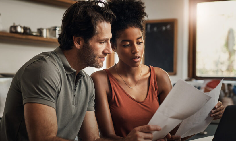 A man and a woman sit together at a kitchen table, looking at documents and a laptop screen with serious expressions. Sunlight comes through the window behind them, and shelves with dishes are visible in the background.