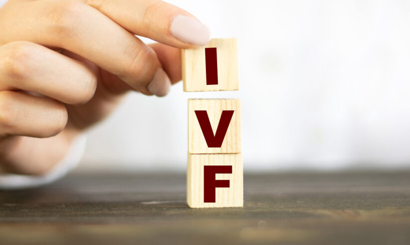 A hand is stacking three wooden blocks with the red letters I, V, and F, forming the abbreviation IVF on a dark surface with a blurred white background.