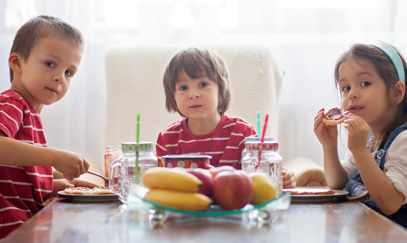 Three young children sit at a table eating together. They all wear red striped shirts. There are plates of food, mason jar drinks with straws, and a fruit bowl with bananas and apples on the table.