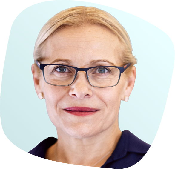 A middle-aged woman with short blonde hair, wearing glasses and a navy blue top, looks directly at the camera against a light blue background.