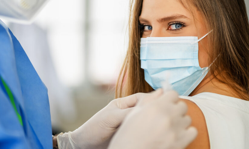 A person wearing a surgical mask receives a vaccine injection in their upper arm from a healthcare worker in protective gloves and a blue gown. The person looks directly at the camera.