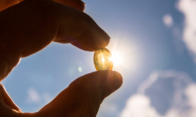A hand holds a translucent capsule up to the sky, with sunlight shining through it and clouds in the background.