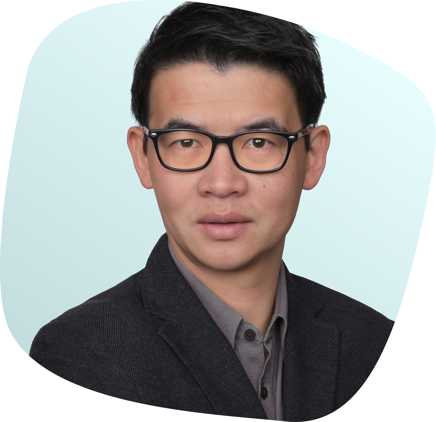 A man with short black hair and glasses wearing a dark blazer and gray shirt poses in front of a light blue background.