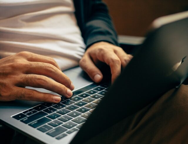 Close-up of a person typing on a laptop. They are wearing a white shirt, dark jacket, and brown pants, and are seated with the laptop resting on their lap. The focus is on their hands and the keyboard.