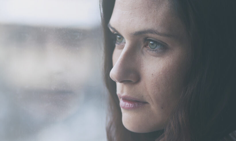 A woman with long brown hair looks pensively out of a window, her face partially reflected in the glass. The mood is contemplative and somber, with soft natural light illuminating her features.