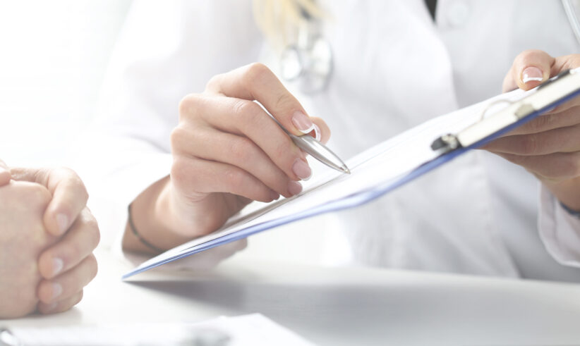 A doctor in a white coat holds a clipboard and pen, discussing medical information with a patient whose hands are folded on the table.