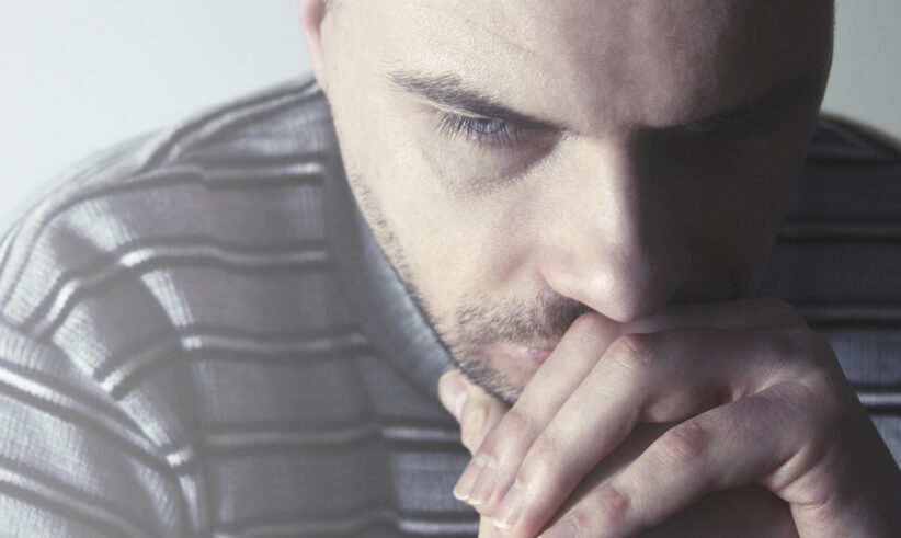 A man in a striped sweater sits with his hands pressed to his mouth, looking downward with a serious and contemplative expression.