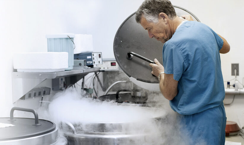 A man in blue scrubs lifts the lid of a large cryogenic storage tank emitting vapor in a laboratory setting with scientific equipment and containers visible.