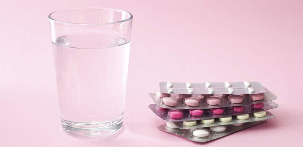 A clear glass of water sits next to four blister packs of various pink and white pills on a light pink background, symbolizing the options and support available, such as egg freezing insurance, for those considering their reproductive health.