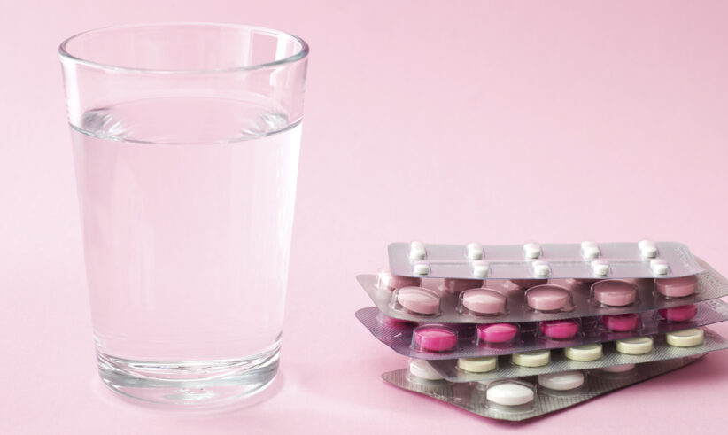 A clear glass of water sits next to four blister packs of various pink and white pills on a light pink background, symbolizing the options and support available, such as egg freezing insurance, for those considering their reproductive health.